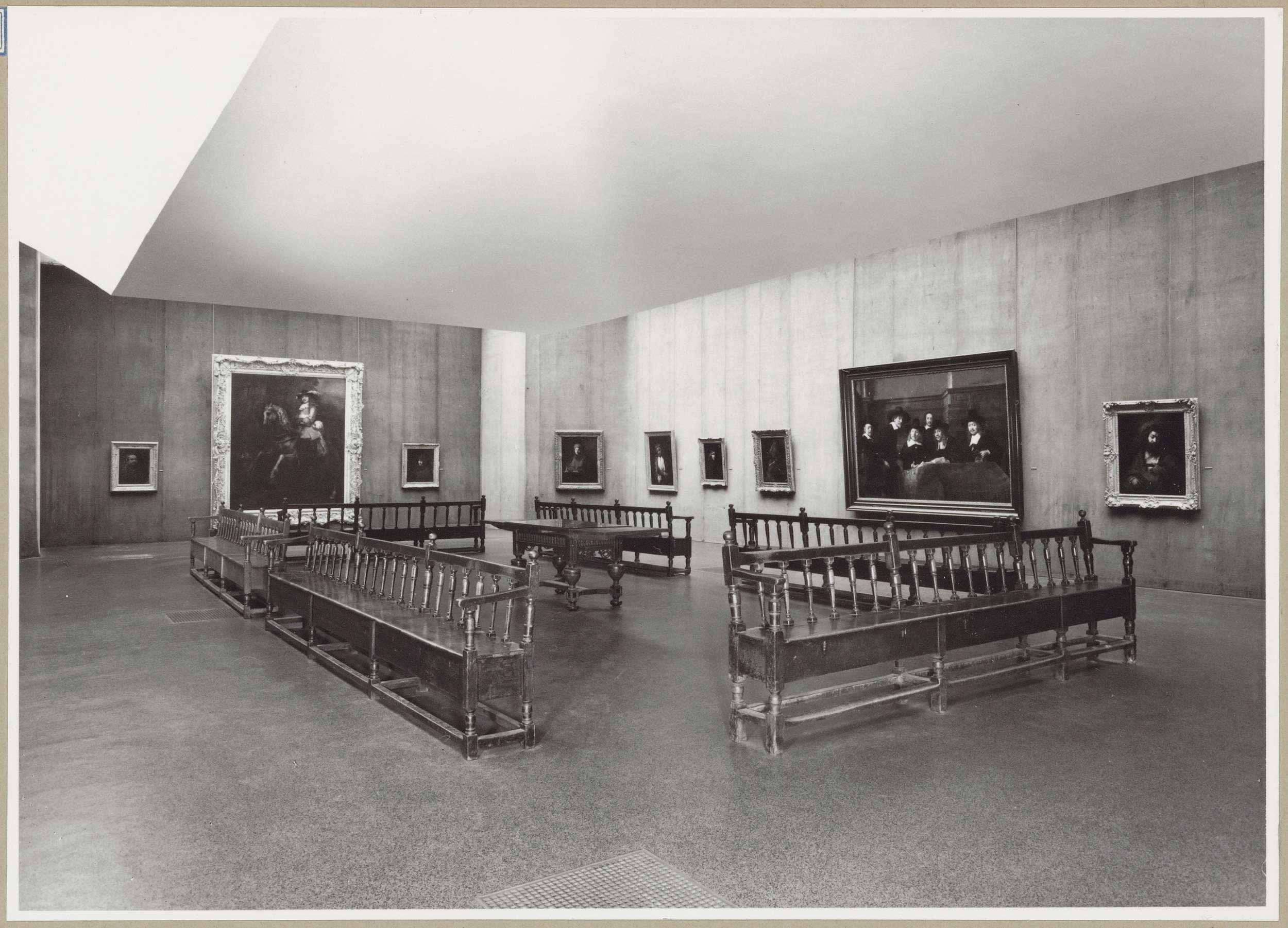 Zaal met negen schilderijen en een tafel te midden van banken